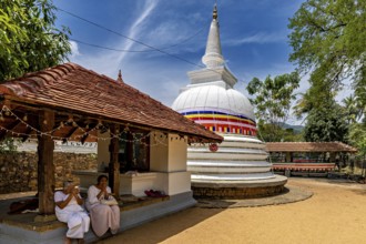 People pray in front of temples and white stupa under a blue sky, surrounded by trees, in a