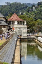 A temple with a pavilion near a river, with people and green surroundings, The Temple of the Tooth