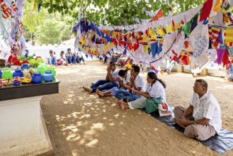 People sitting praying under colorful flags in the shade of a tree on sandy ground, pilgrims to the