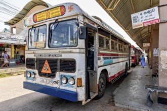 An Ashok Leyland bus at a busy bus stop in Sri Lanka, surrounded by signs and passers-by, bus