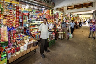 A man stands in a well-stocked shop with numerous colorful foods and drinks, dealers in the market