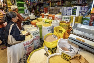 Market customers buy rice from large bags at a well-stocked stand, dealers in the market halls of