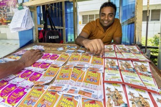 A man smiling behind a sales desk with colorful lottery tickets and tickets, dealer in the market