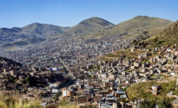 A city nestles on mountain slopes under clear blue skies, lined with green hills and dense