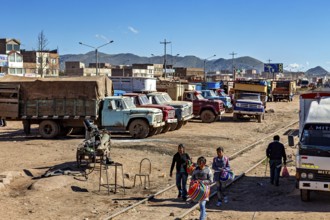 Lively market with people and colorful trucks against a mountain backdrop and clear skies, people