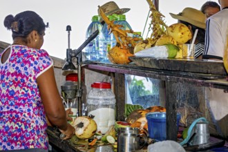 Woman at a market stall cuts coconuts surrounded by tropical fruits and everyday objects,