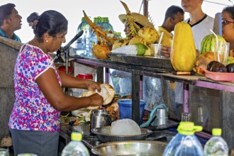 Woman prepares coconuts at a busy market stall surrounded by people and tropical produce,