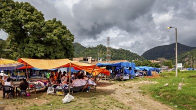 Open-air market with various stalls under orange and blue covers in front of mountains, The market