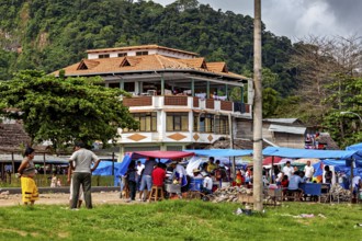 Large house in the background of a busy market square with people and mountains in the distance,