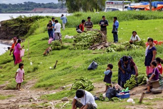 People sell bananas on a riverbank in a rural area. The scene depicts a communal and bustling