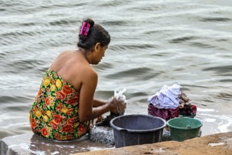 A woman is sitting by the river washing clothes with several buckets, a woman is washing at a lake
