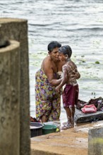 A woman washes a child at the river, both laugh and wear colorful clothes, a woman washes at a lake