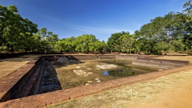 An empty brick water pool in a historic setting with green trees, the temples, dagobas, and pagodas