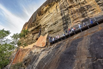 People climb a steep staircase on a rock wall under a blue sky, The Lion Rock near Sigiriya in Sri