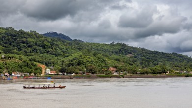 A boat moves on a river in front of a wooded hill and under a cloudy sky, people on boats near