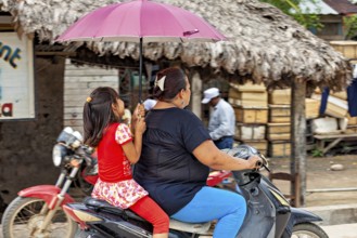 A woman and a child ride on a moped under a red umbrella in a tropical setting, A woman and child