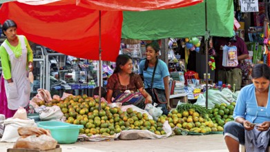 Two laughing woman sit in front of market stalls full of mangoes in a lively atmosphere, The