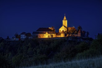 An illuminated castle is perched on a hill, surrounded by dark countryside and a deep blue night