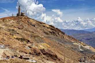 Rough mountain terrain with sparse vegetation and a transmission mast on the summit under a cloudy