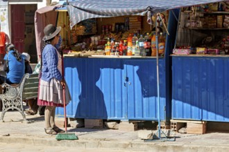 Woman in traditional dress sweeps in front of a blue market stall full of colourful bottles and