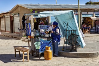 Street snack bar under a blue sunshade, woman serving customers next to a table and gas canister in