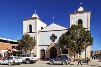 Church with two towers and cross in a busy street, surrounded by cars and people under a clear blue