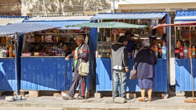 Street market with a blue stall, people buy colourful bottles and products under a blue sky, small