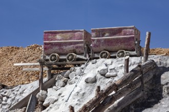 Two rusty mine cars on a wooden scaffold, surrounded by piles of rocks in a sunny mining