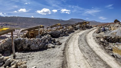 Dusty path with piles of stones next to a mountainous landscape under a blue sky with clouds, The