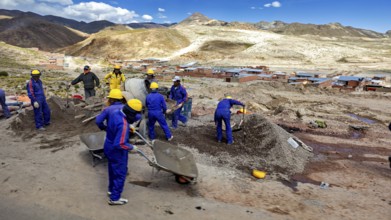 Workers in blue work clothes shovel earth on a sunny hilly landscape, surrounded by mountains and
