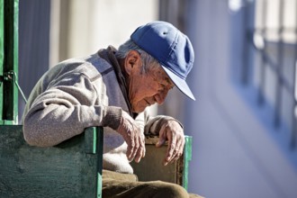 Elderly man in cardigan and blue cap sits relaxed, surrounded by sunshine and tranquillity, show