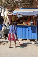 Woman in traditional dress sweeping in front of a blue stall on a busy street in the sunshine,