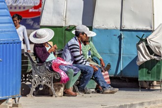 People with hats sitting on a bench in the sun at a market stall, people of the city of Uyuni in