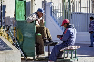 Shoe shiner concentrates on a man sitting on a green bench outdoors, show shiner in the city of