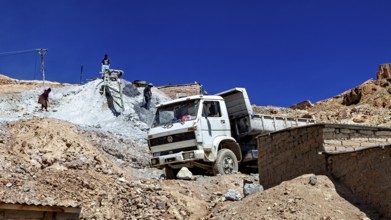 A lorry unloads gravel at a construction site in a mountainous desert landscape under a clear sky,