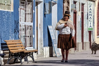 Woman with a traditional hat stands in front of a blue house wall in the sun, people of the city of