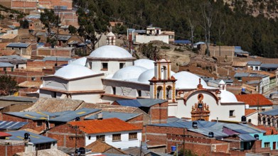 A city view with domes of a church in the foreground, surrounded by brick houses in a mountainous