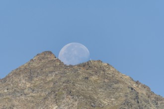 Majestic mountain peak stands tall against vibrant blue sky as large moon rises just behind it. The