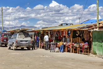 Various market stalls with visitors and cars, lively rural atmosphere under a cloudy sky, market