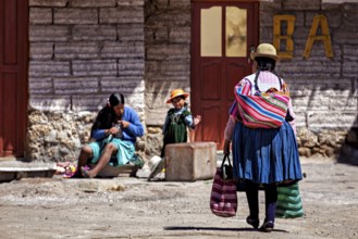 A woman approaches a small group in which a child is playing and another woman is sitting,