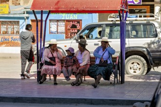 People in traditional dress sit on a bench in an urban environment as a car drives past, people at