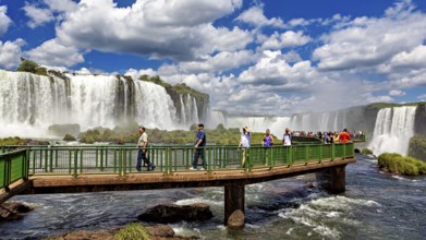 People walk on a bridge in front of an impressive waterfall under a blue sky, The Iguazu Falls