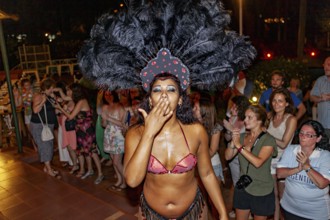 Woman in exotic costume with feather headdress gives kisses to the crowd, samba dancers to
