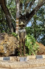 Bronze statue of a man with a hat and suit, surrounded by trees and rocks in a natural setting,