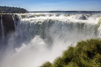 Plunging water of the waterfall with dense foam, surrounded by green vegetation under a blue sky,