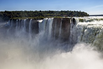 Thick mist rises from the mighty waterfall, flanked by green forest under a blue sky, The Iguazu