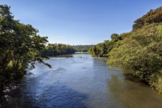A calm river flows through a forested landscape under a clear blue sky, The Iguazu River between