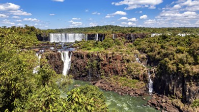 Large waterfalls flow over steep cliffs, surrounded by lush greenery and a blue sky with clouds,
