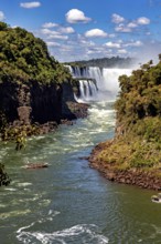 Dramatic waterfalls pour into a river, surrounded by dense vegetation and under a brilliant sky,