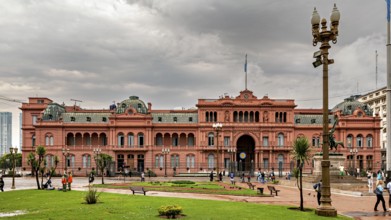 A historic, pink colonial-style building with a cloudy sky and palm trees in the foreground, the